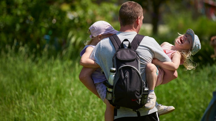 A man facing away from the camera with a backpack on carrying two small children who are both smiling. It is a sunny summer day.